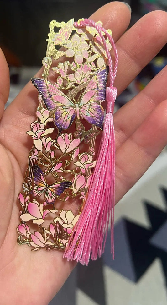 Metallinen kirjanmerkki kädessä: vaaleanpunaiset magnoliat, violetit perhoset ja vaaleanpunainen tupsu